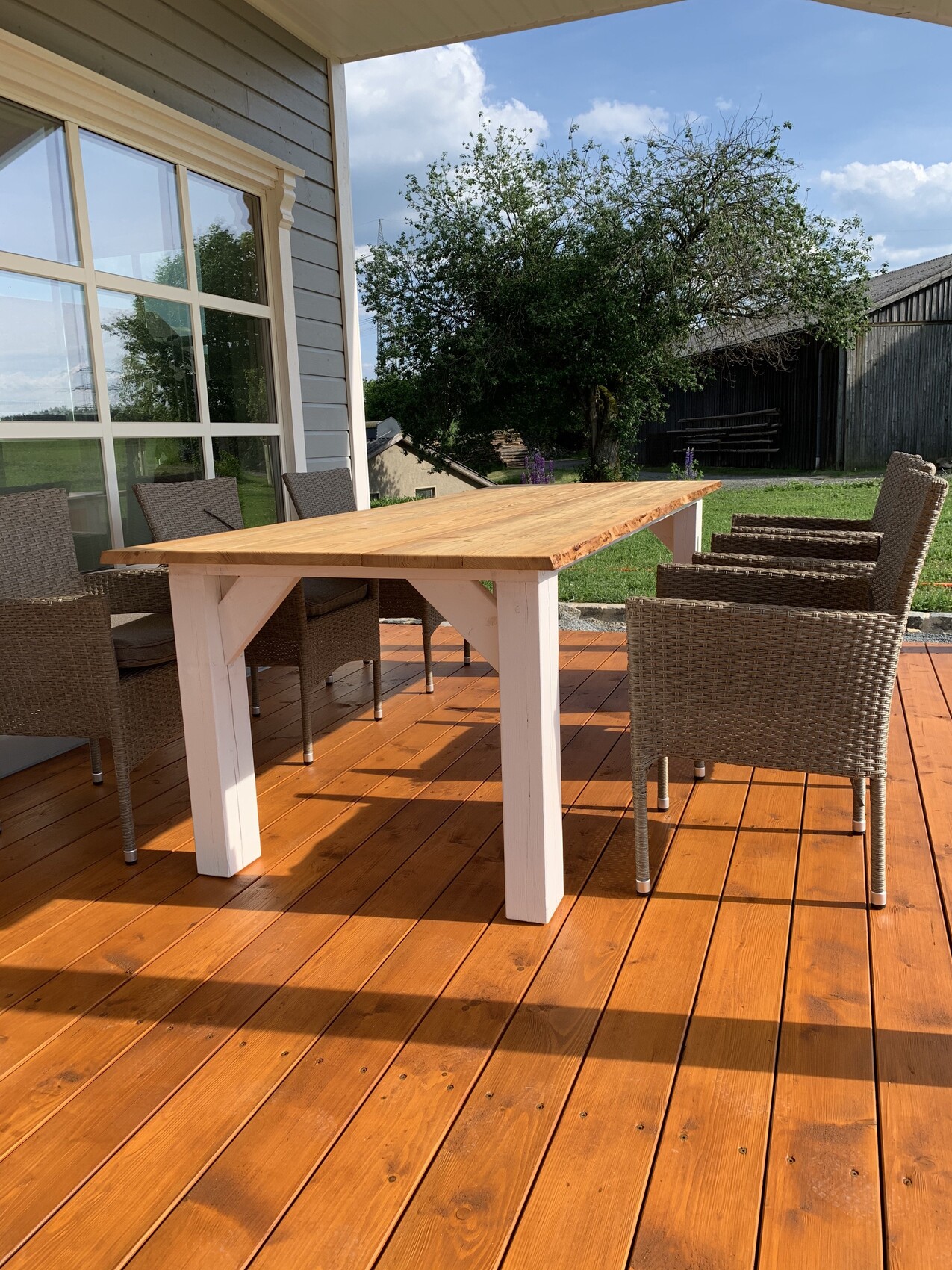Holzdeck mit großem Esstisch und Korbstühlen vor einem Haus mit großen Fenstern; Baum im Hintergrund