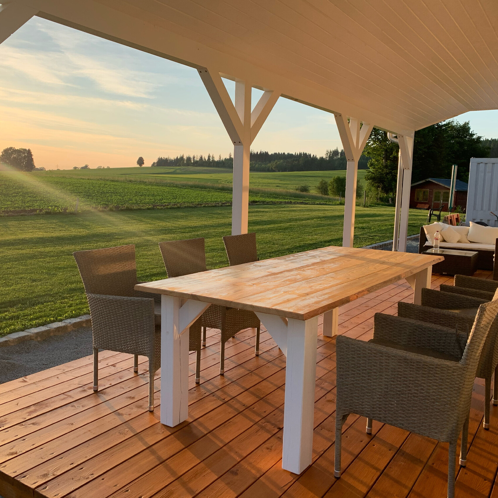 Großer Holztisch mit Korbsesseln auf der Veranda, Blick über grüne Felder bei Sonnenuntergang