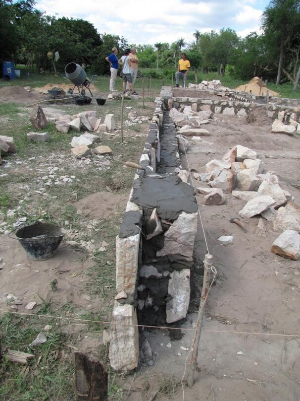 Construction workers building a stone wall with mortar in a pit; other workers in the background.