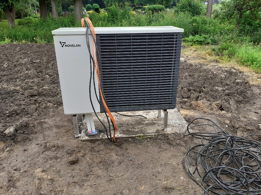 Outdoor unit of an air conditioning system (condenser) with NOVELAN logo on concrete foundation; cables next to it.