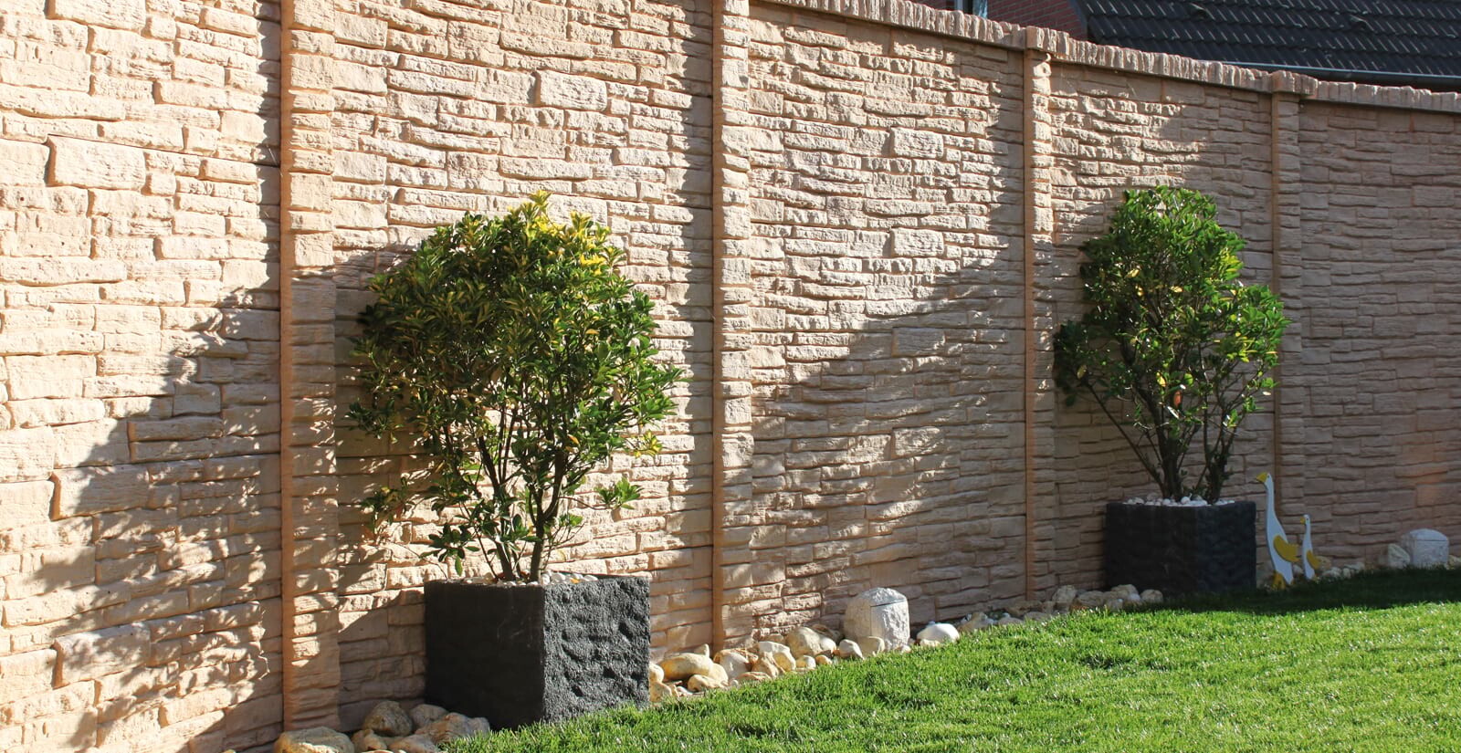 Two green shrubs in black plant containers in front of a multicolored stone wall, grass in front