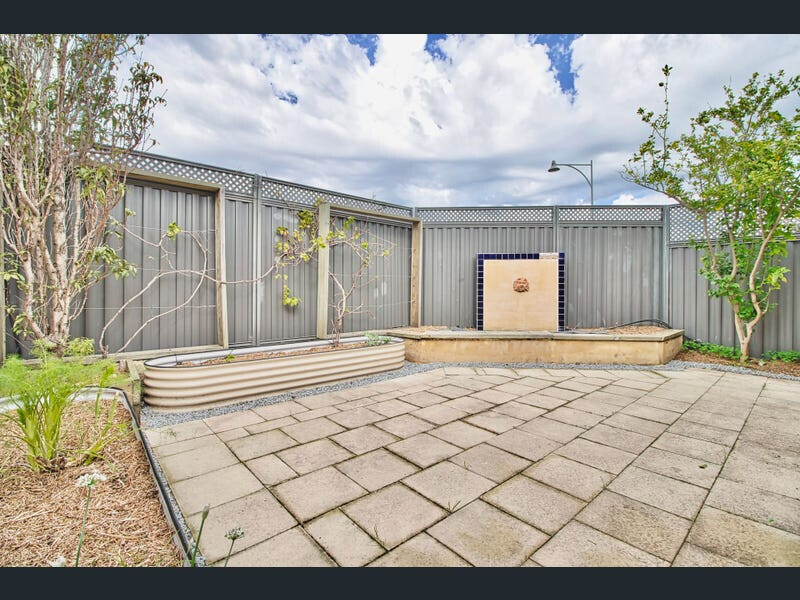 Garden courtyard with fountain, gray fence, planted flower beds, and paved ground.