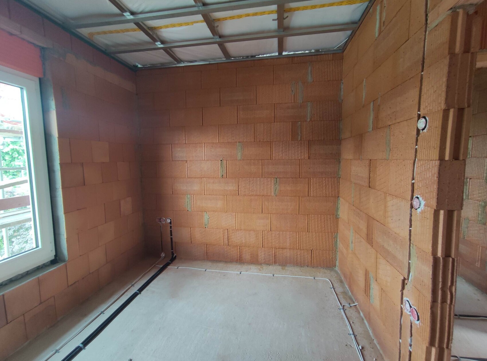 Shell construction room with red brick walls, window on the left, visible cables on the floor, open ceiling.