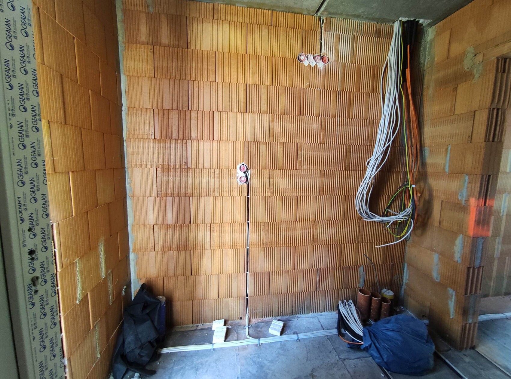 Interior of a construction site with brick wall, visible cable bundles, pipes and components on the floor.