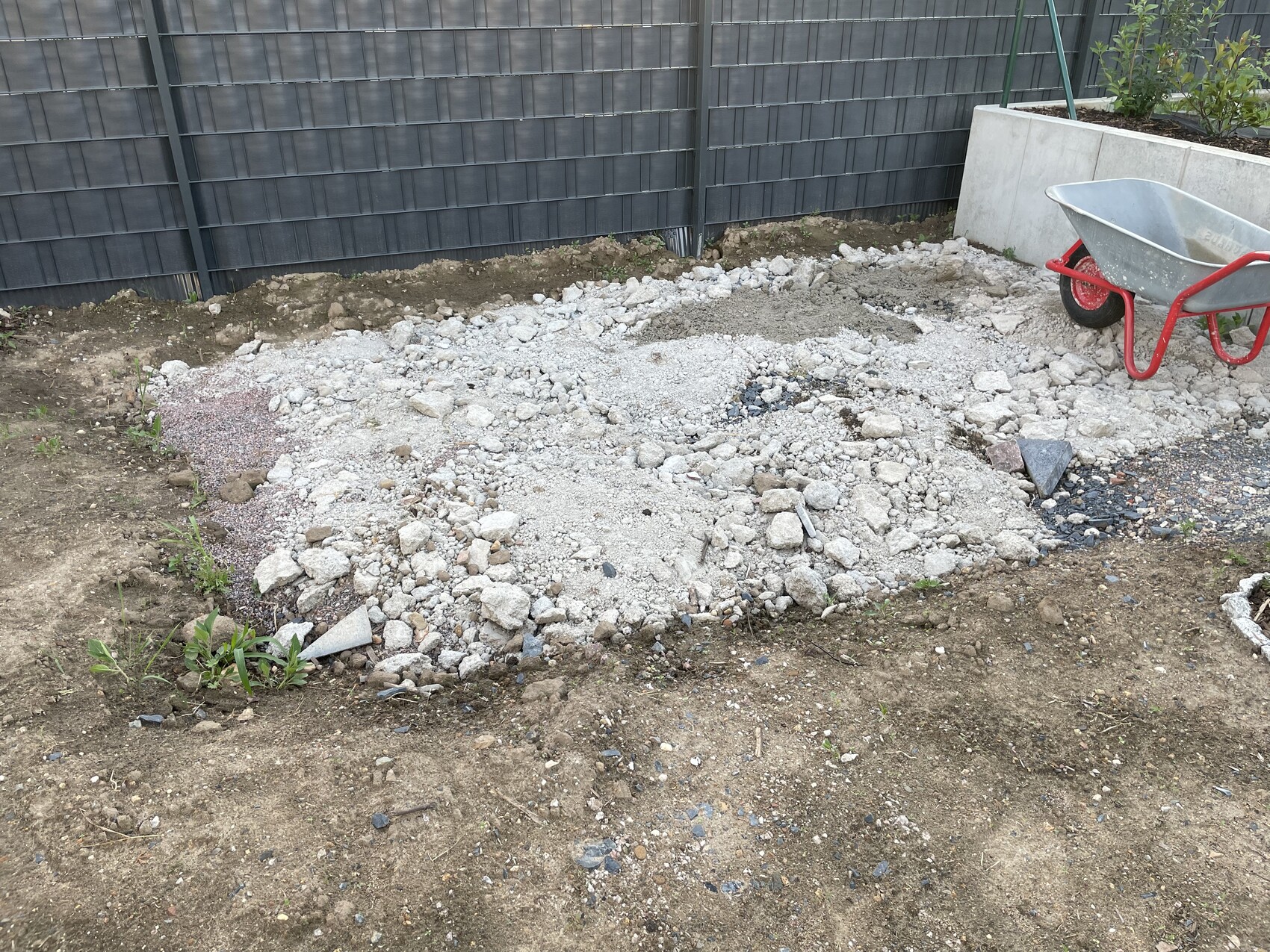 Construction site in the garden: gravel, rubble, and soil excavation, red wheelbarrow next to dark fence.