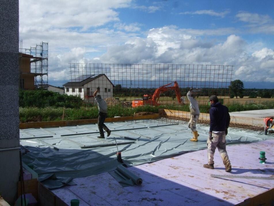 Construction worker laying foil on a building site; excavator in the background.