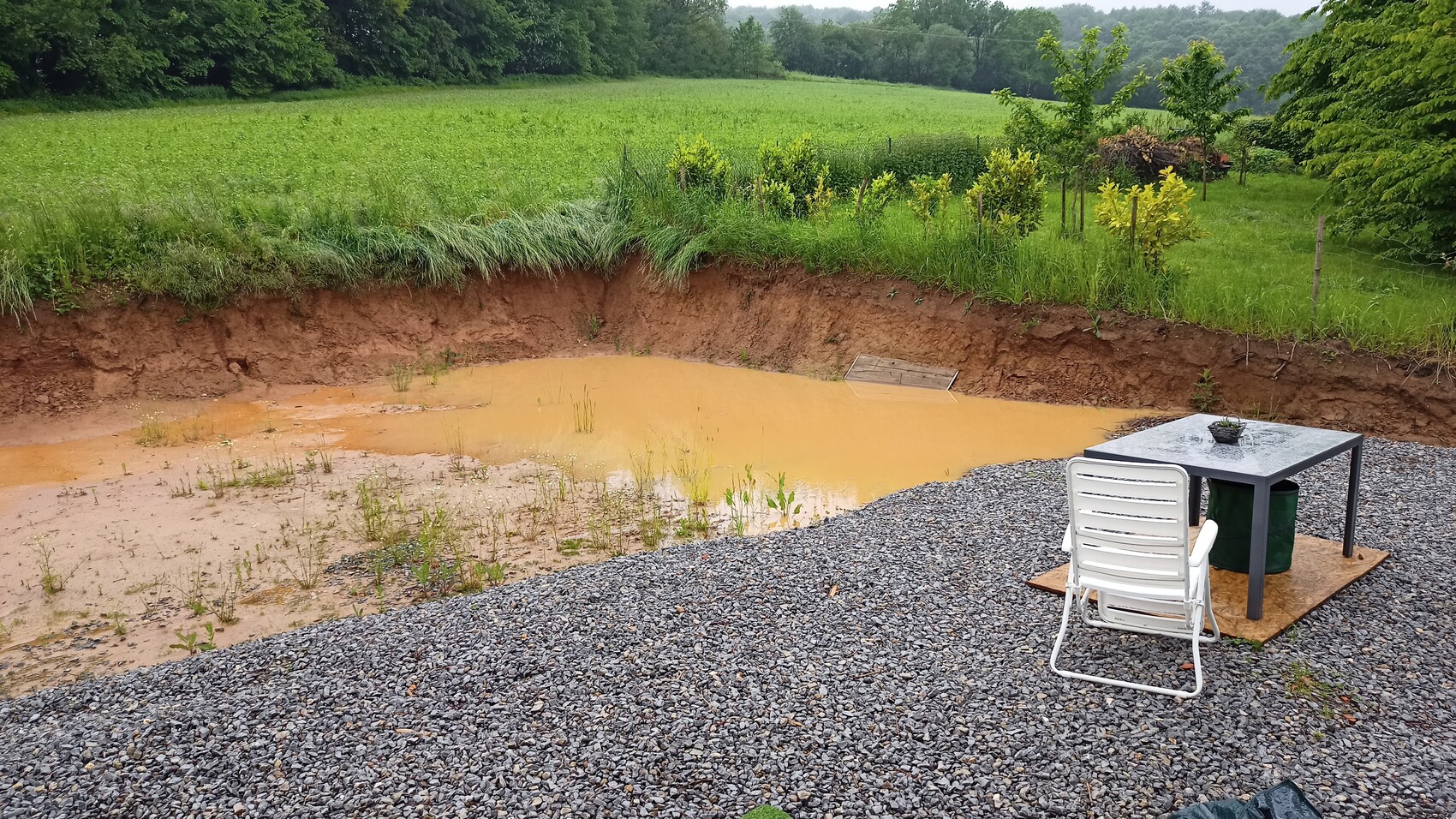 Braunes Schlammloch in einer Grube, daneben Tisch und Stuhl auf Kiesfläche, Gartenlandschaft.