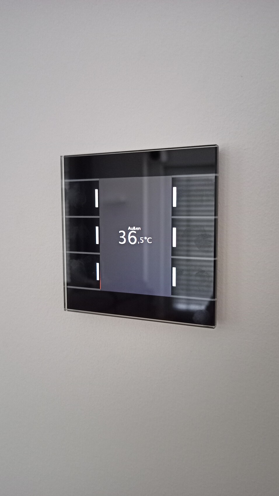 Wall-mounted smart thermostat with glass display shows outdoor temperature 36.5°C.