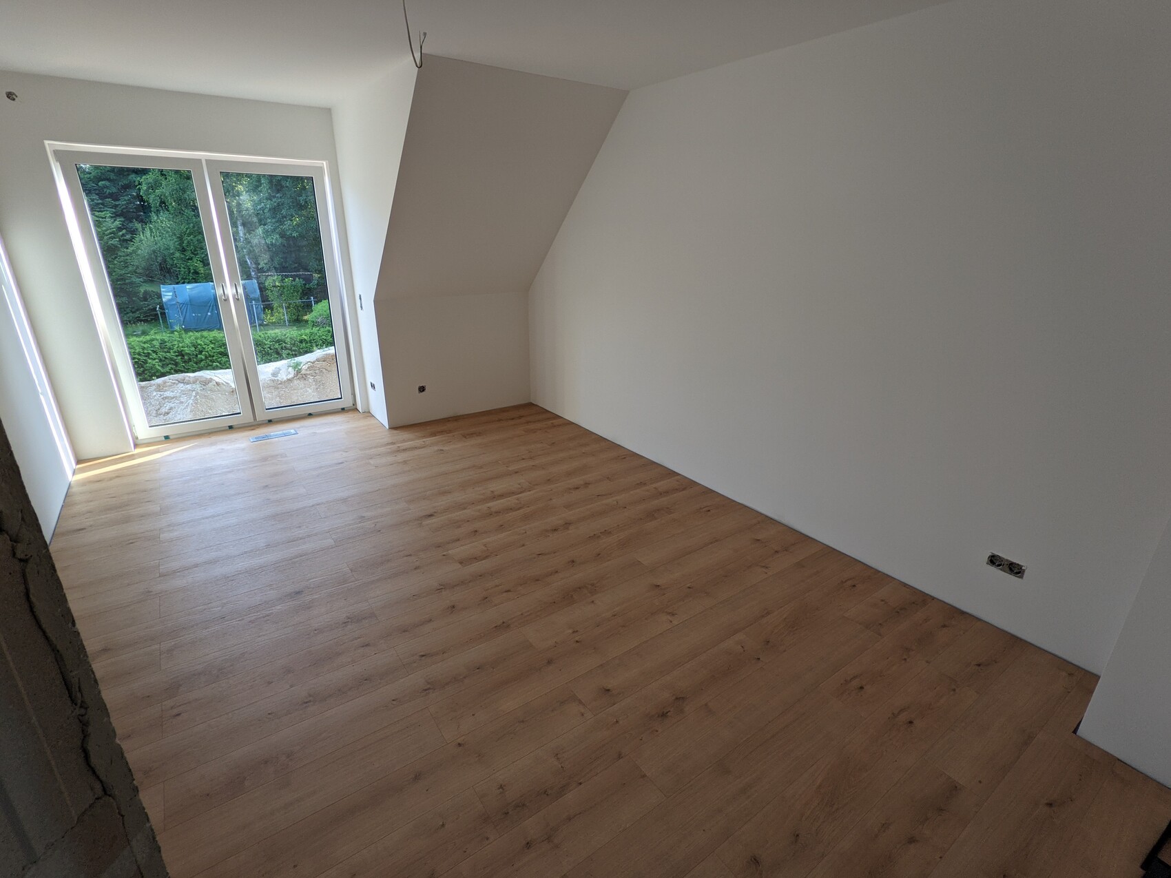 Bright, unfurnished room with wooden floor; glass sliding door to the garden and green outdoor area.