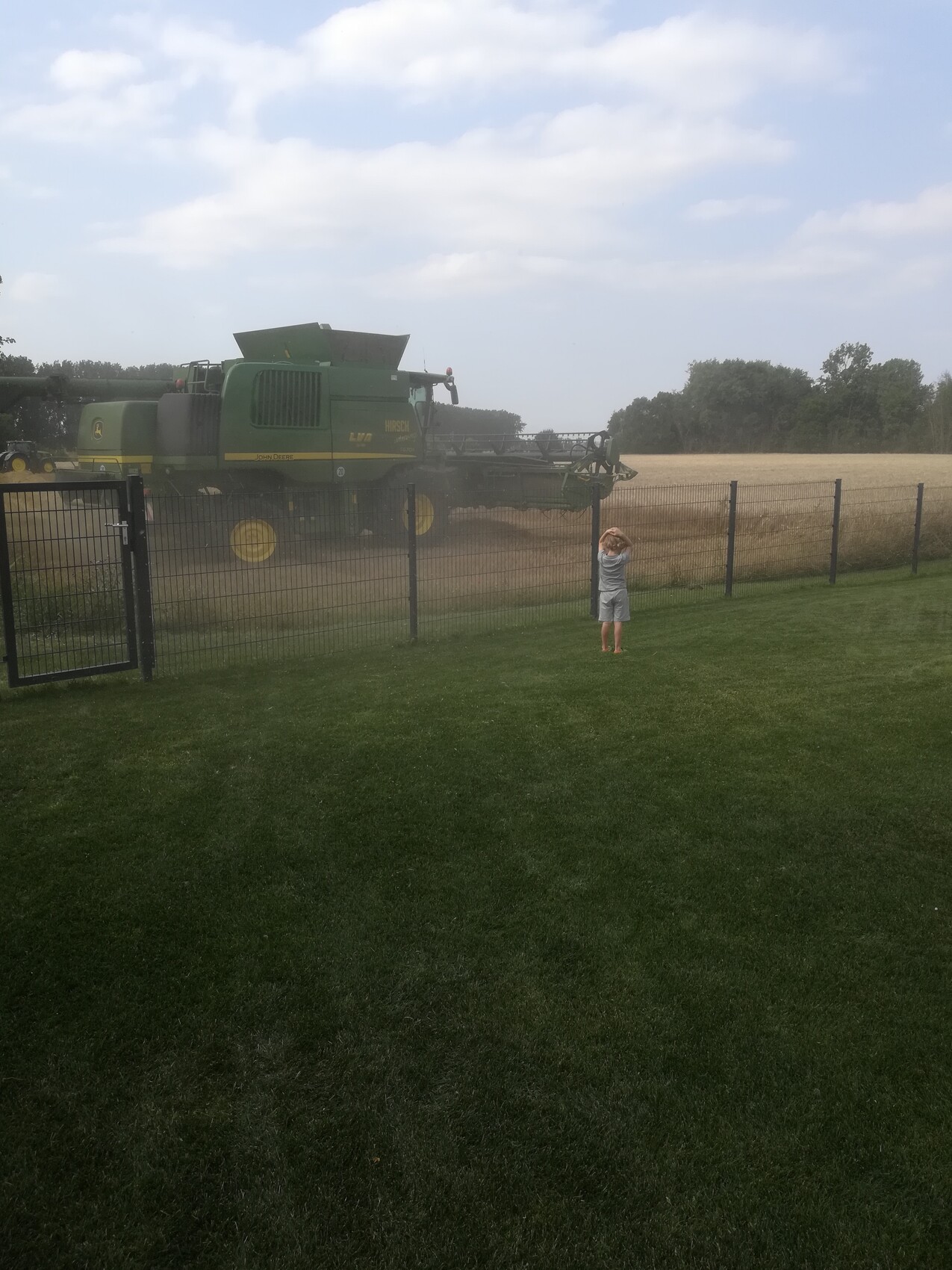 Small child in front of a fence watching a large green combine harvester on a dusty field.