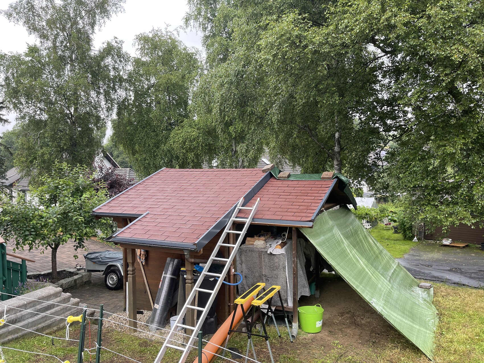 Small wooden house with red roof, ladder in front, green tarp as a cover in the garden.