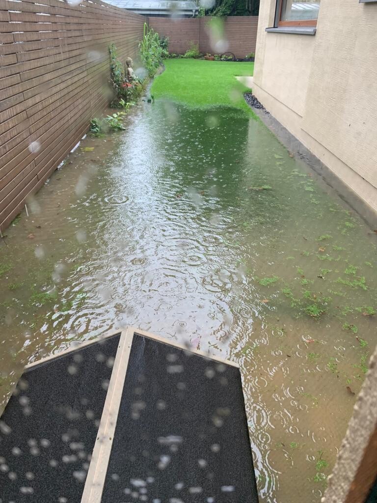Water-flooded narrow garden between wooden slat fence and house wall; water covering the ground.