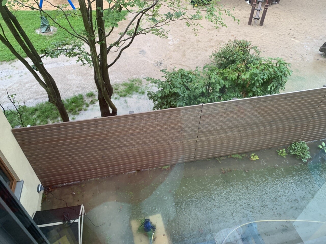 View from inside on a brown slat fence, flooding in the yard due to rain, trees in the background.
