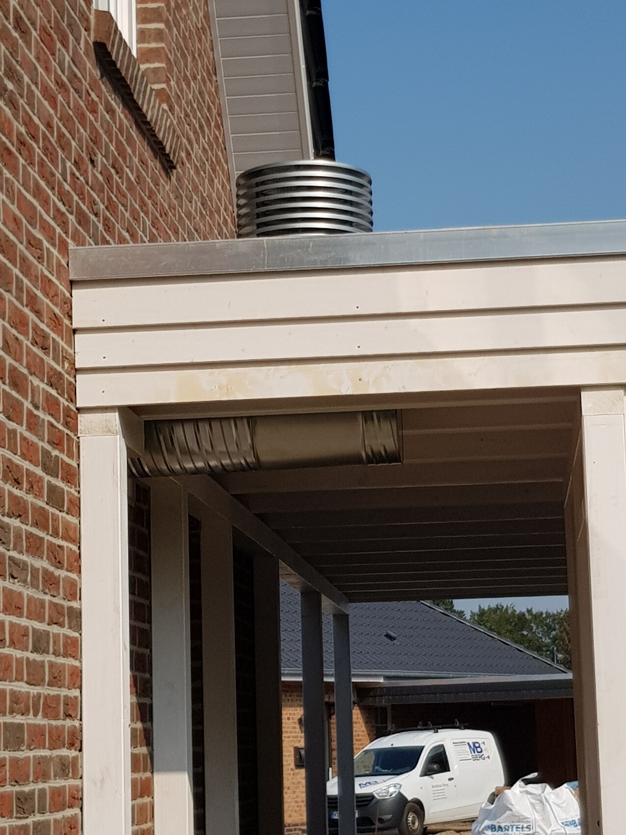 Round metal ventilation pipe on flat roof next to carport brick wall with blue sky on the left.