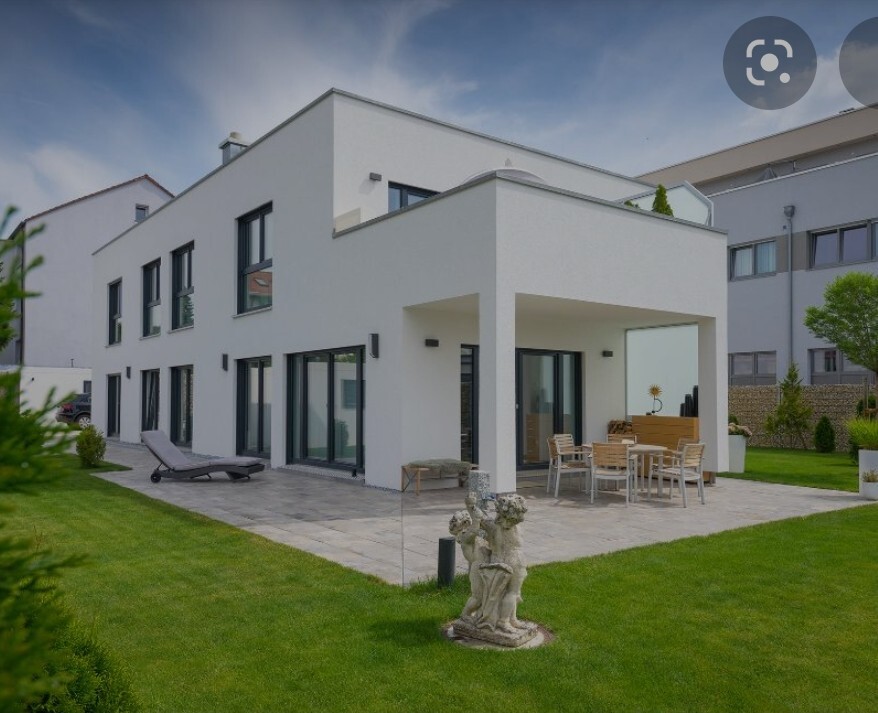 Modern white villa with terrace, outdoor dining area and lounge chairs; sculpture on the lawn.
