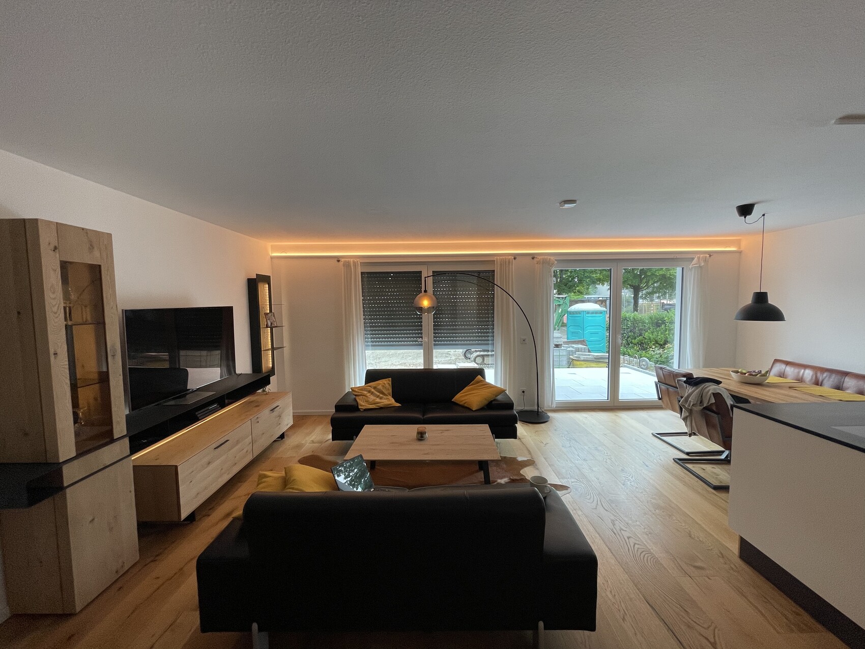 Wohnzimmer mit schwarzem Sofa, Holzboden TV-Schrank, Esstisch rechts, Glas-Schiebetüren nach außen.