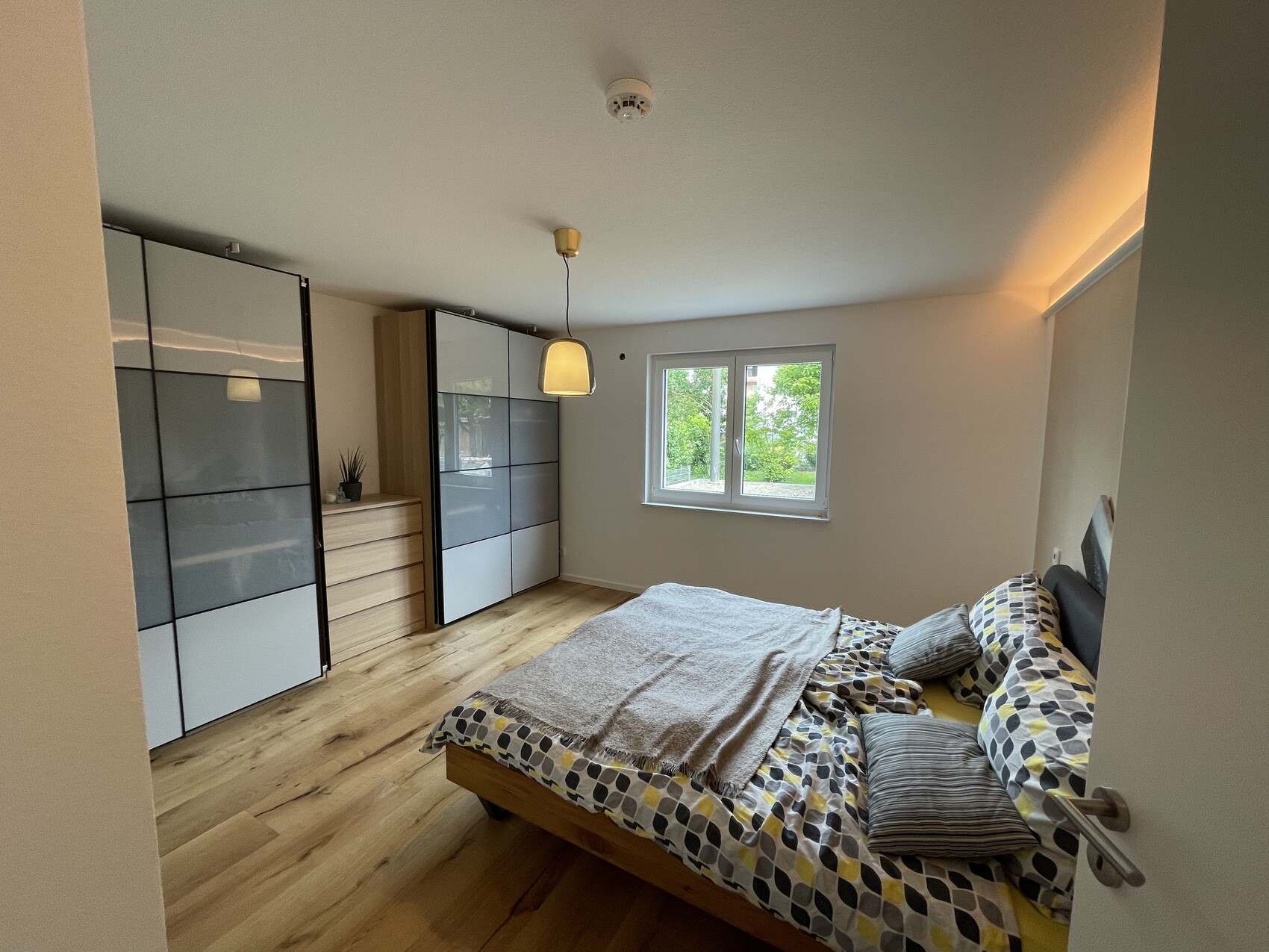 Schlafzimmer mit Bett, gerahmten Kleiderschränken, Kommode, Holzboden und Fenster mit Grünblick.