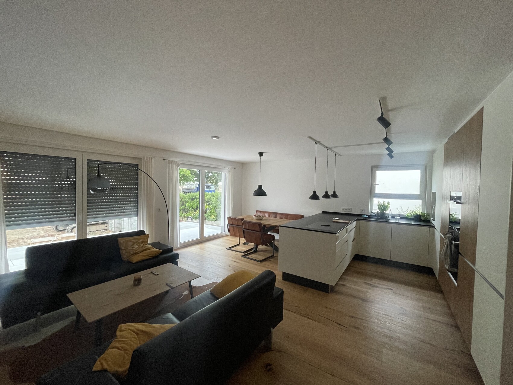 Offene Wohnküche mit Insel, Esstisch, schwarze Sofas, Glastüren zum Garten und Pflanzen.