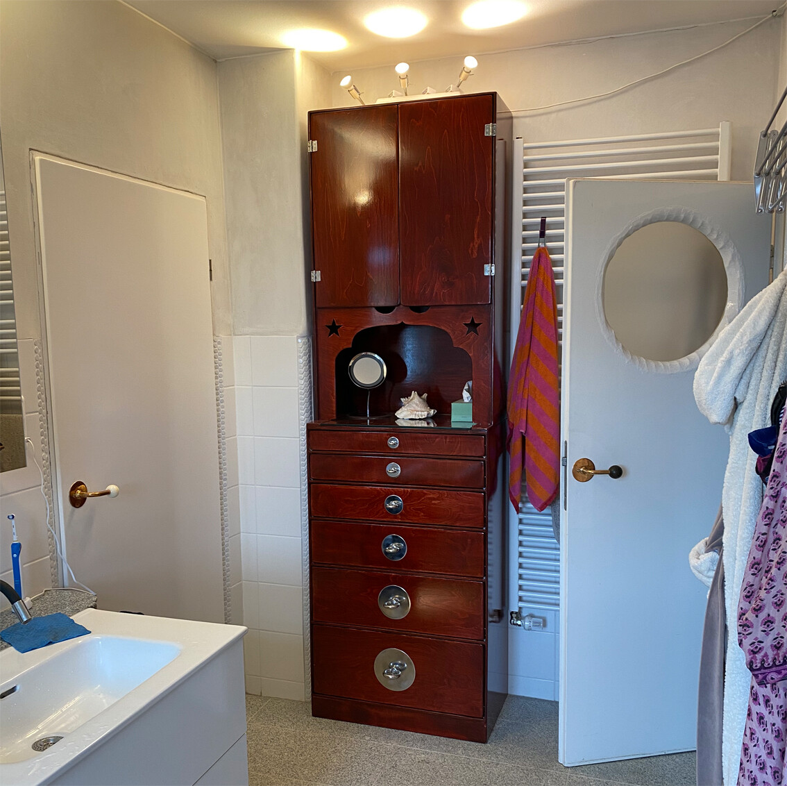 Bathroom: red wooden cabinet tower, sink on the left, mirror, door on the right with round window.
