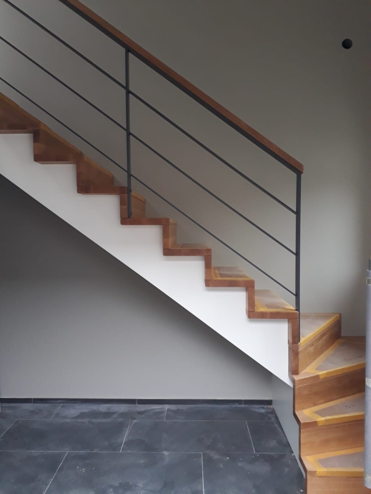 Modern wooden staircase with black metal railing in front of a gray wall and dark tiled floor.