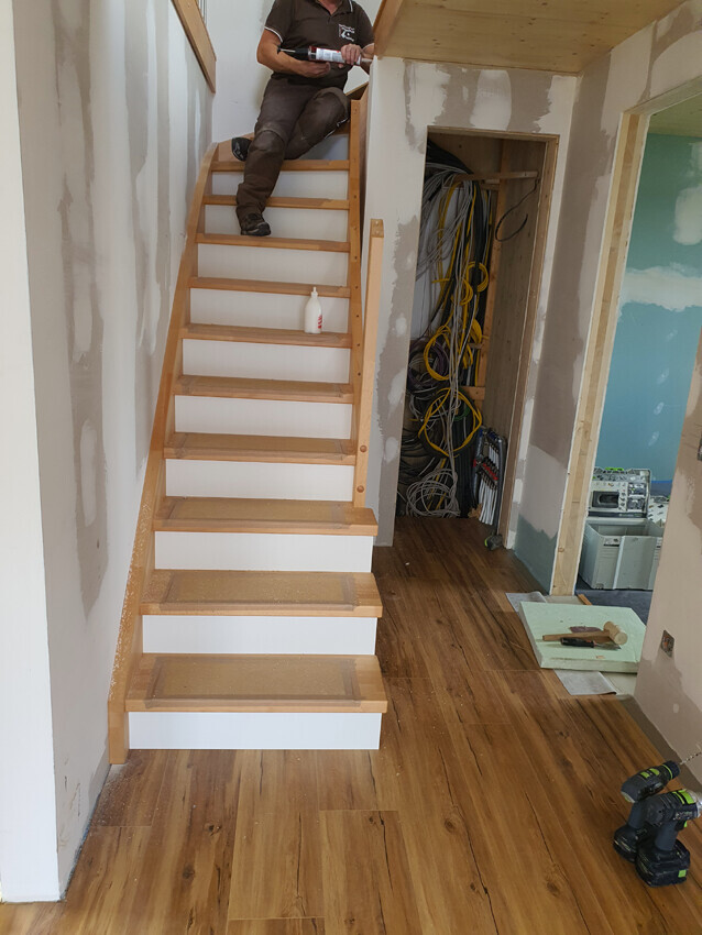 Worker on wooden staircase during construction, tangled cables behind open door, drill on the floor.