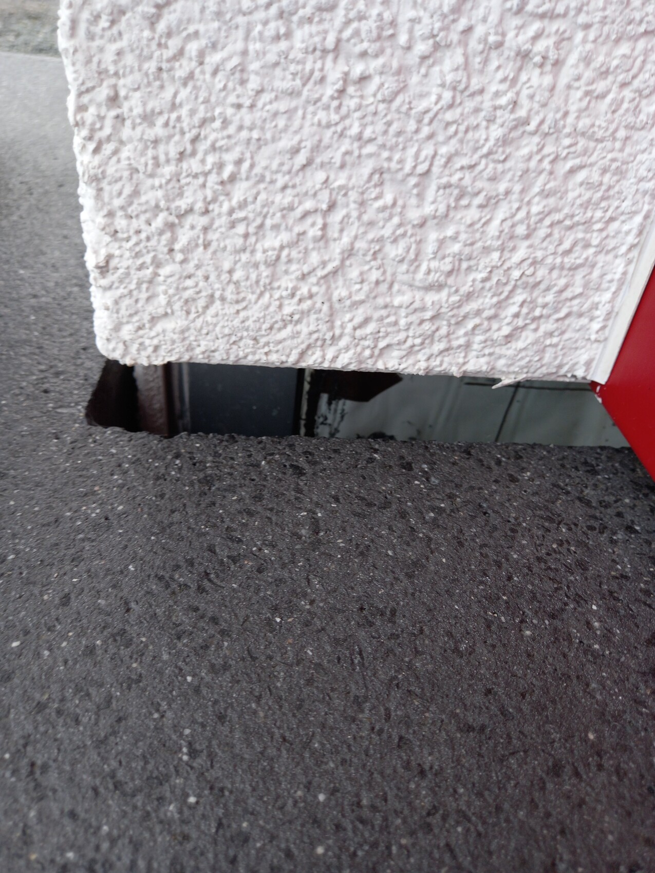 Close-up of a rough white wall above a hole in the asphalt, with a red metal edge visible at the bottom