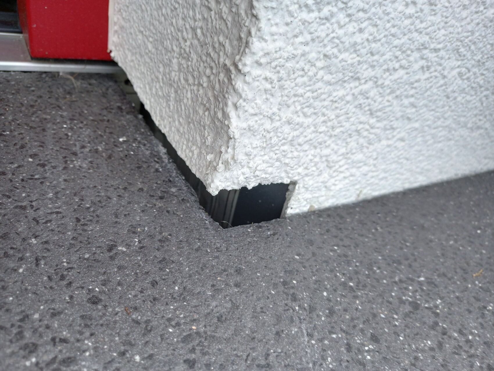 Coarse white exterior wall with a rectangular opening at the bottom, dark interior visible, asphalt in front.