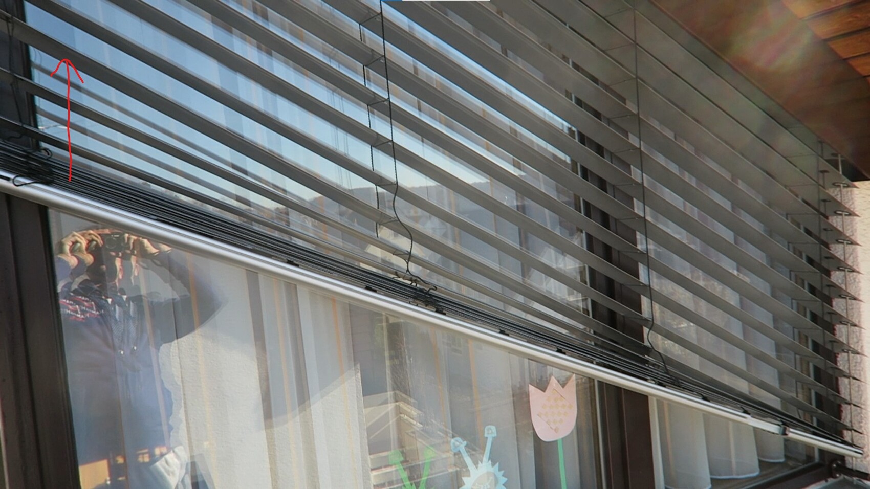 Window with narrow horizontal blinds; reflection of a person holding a camera.