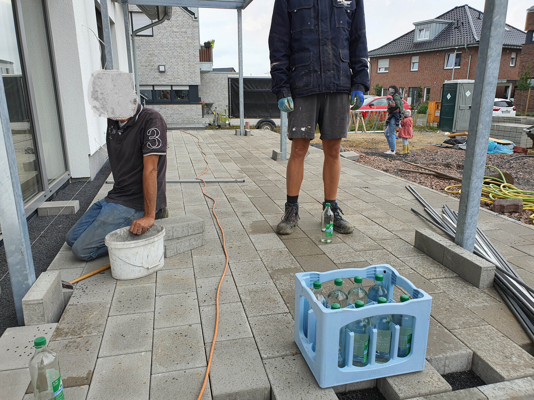 Zwei Bauarbeiter verlegen Pflastersteine auf einer Terrasse; Eimer, Getränkekisten.