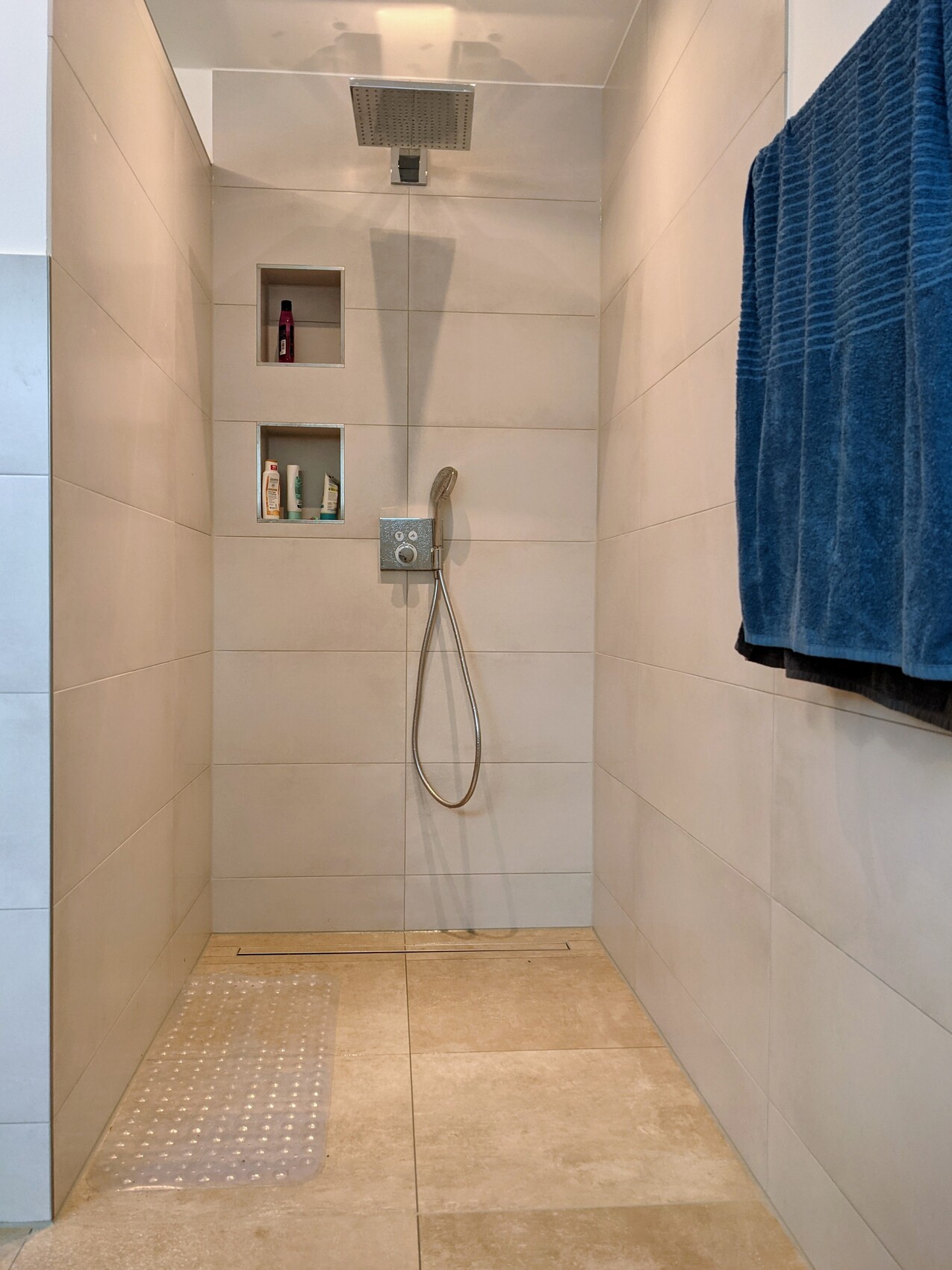 Moderne Walk-In-Dusche mit Regenkopf, Handbrause, zwei Ablagen und blauen Handtüchern.