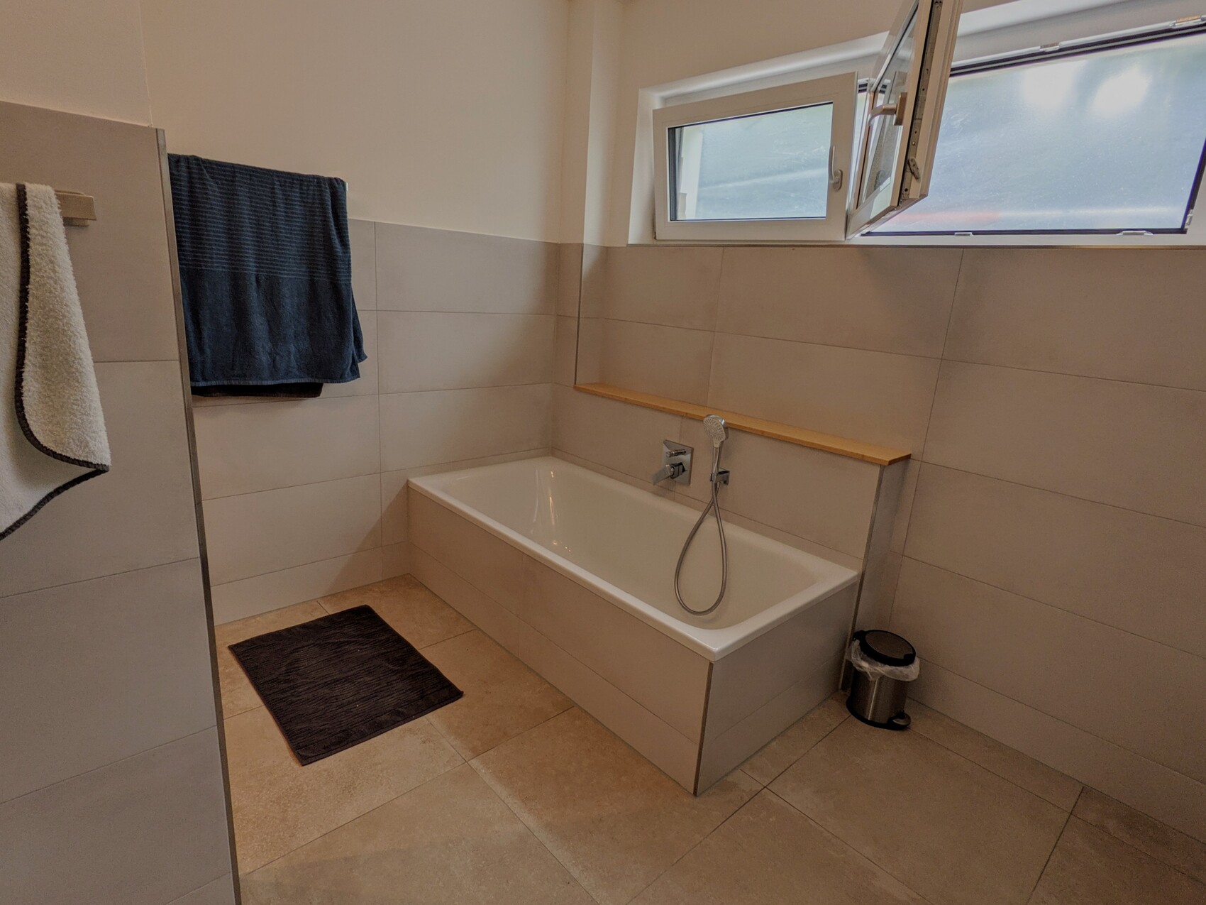 Bathroom with white bathtub, window above, handheld shower, dark bath mat.