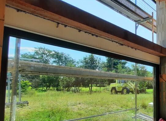 View through an open window: scaffolding, green meadow and a yellow construction vehicle in the background.