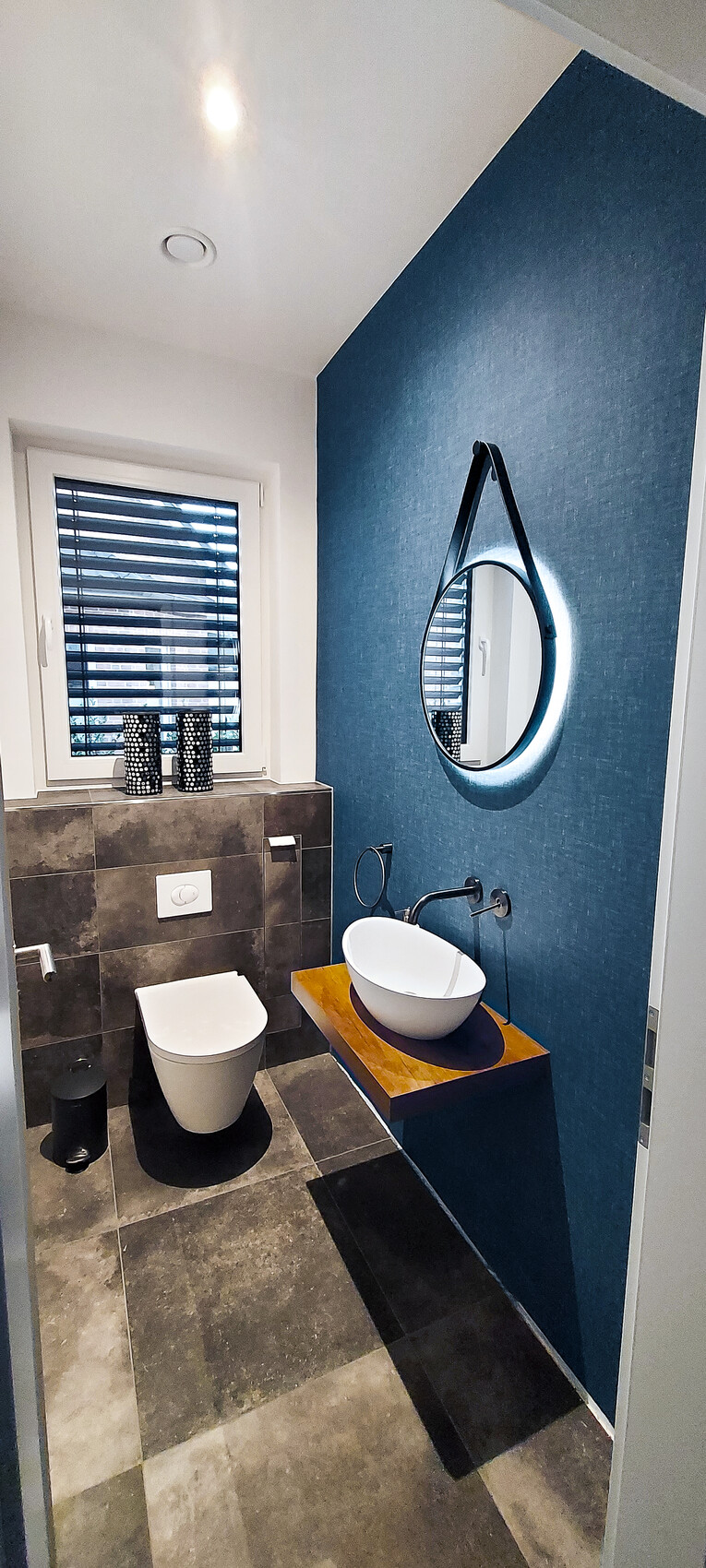 Modern bathroom: blue accent wall, round illuminated mirror, sink on wooden countertop, toilet.