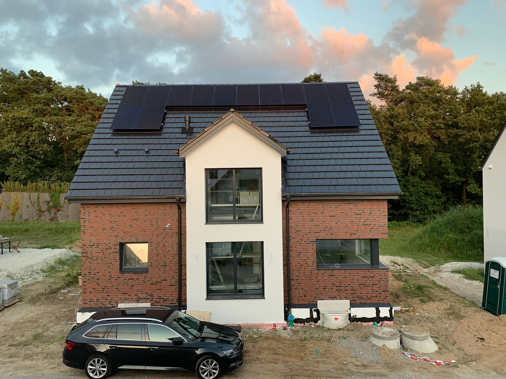 Two-story house with dark tiled roof, solar panels, red brick, and black car in front.
