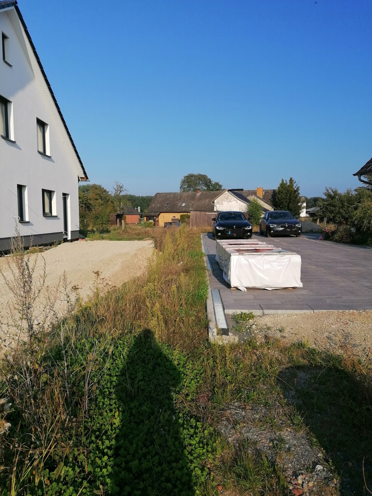 Modern white facade on the left, paved driveway with wrapped block, two cars, blue sky.