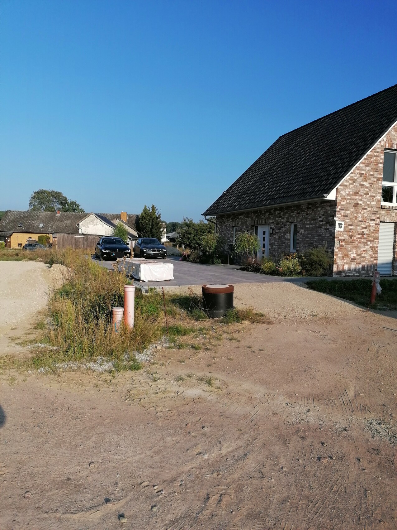 Brick house with dark roof on the right, unpaved driveway, construction items, cars