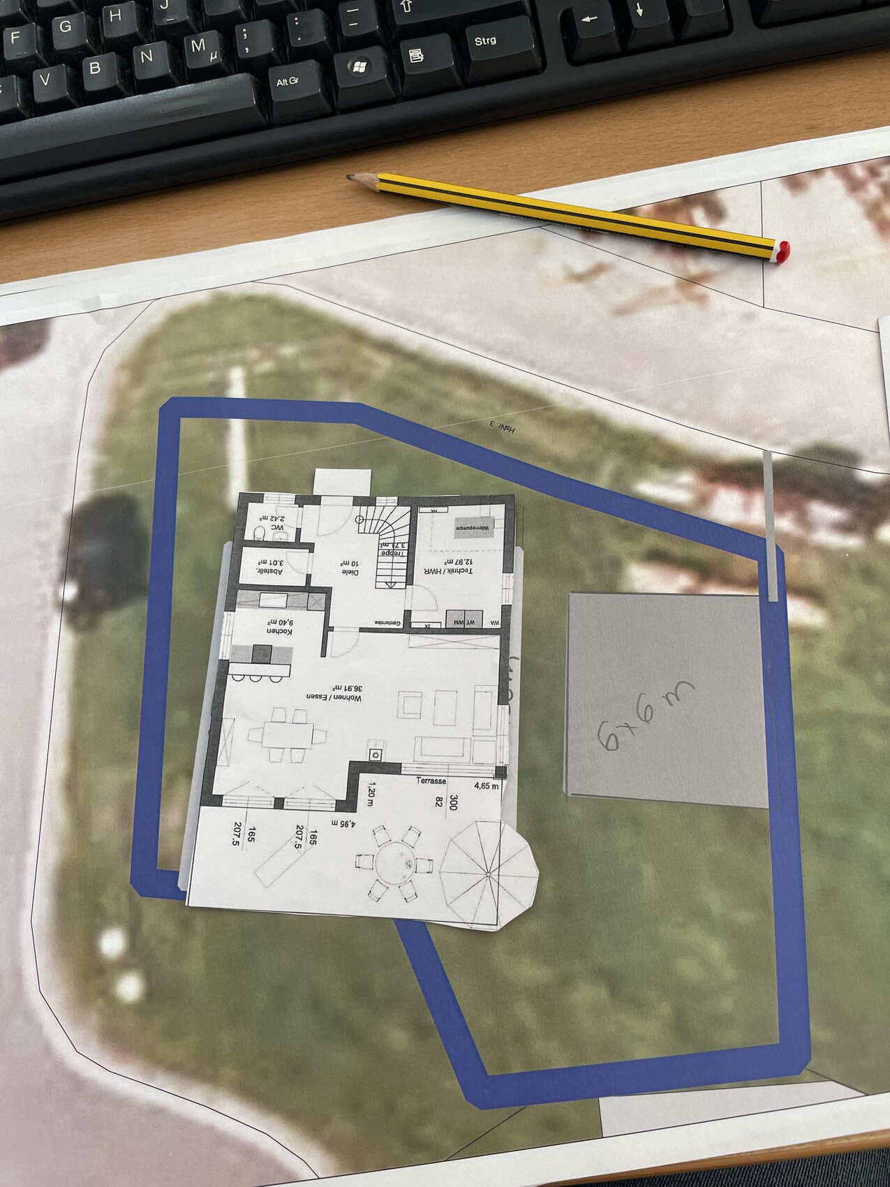 Paper model of a house floor plan on a plot map with terrace; pencil beside it.