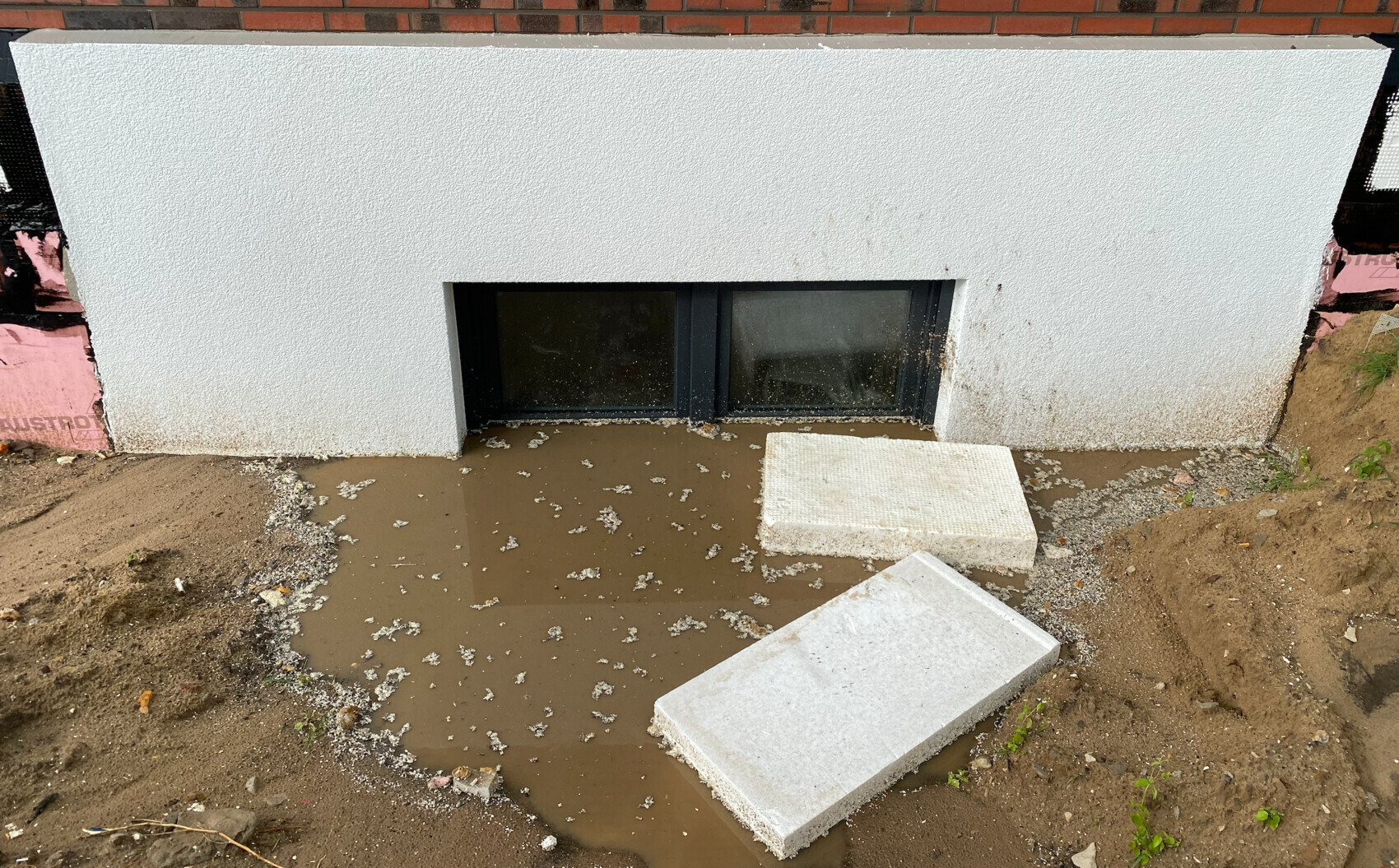 Basement window in white, rough plaster facade; in front, a mud puddle and two loose concrete slabs.