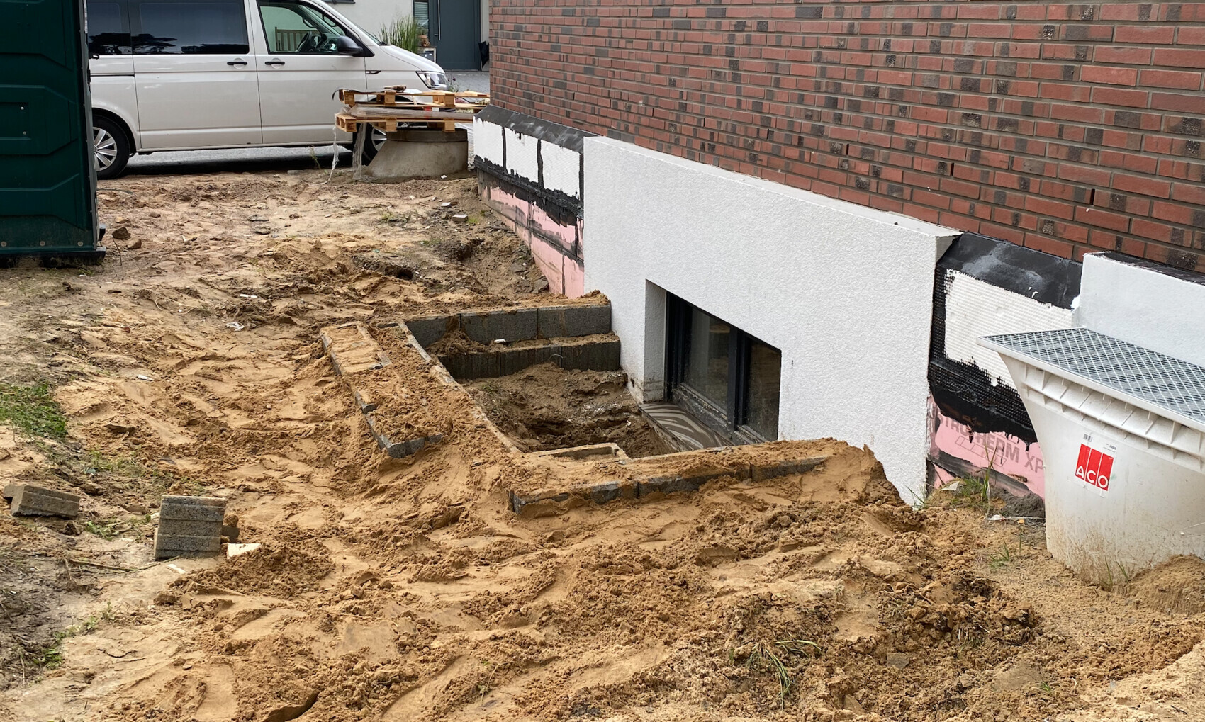 Construction site at the house: open basement window, earthworks, sand piles, pallets on the left.