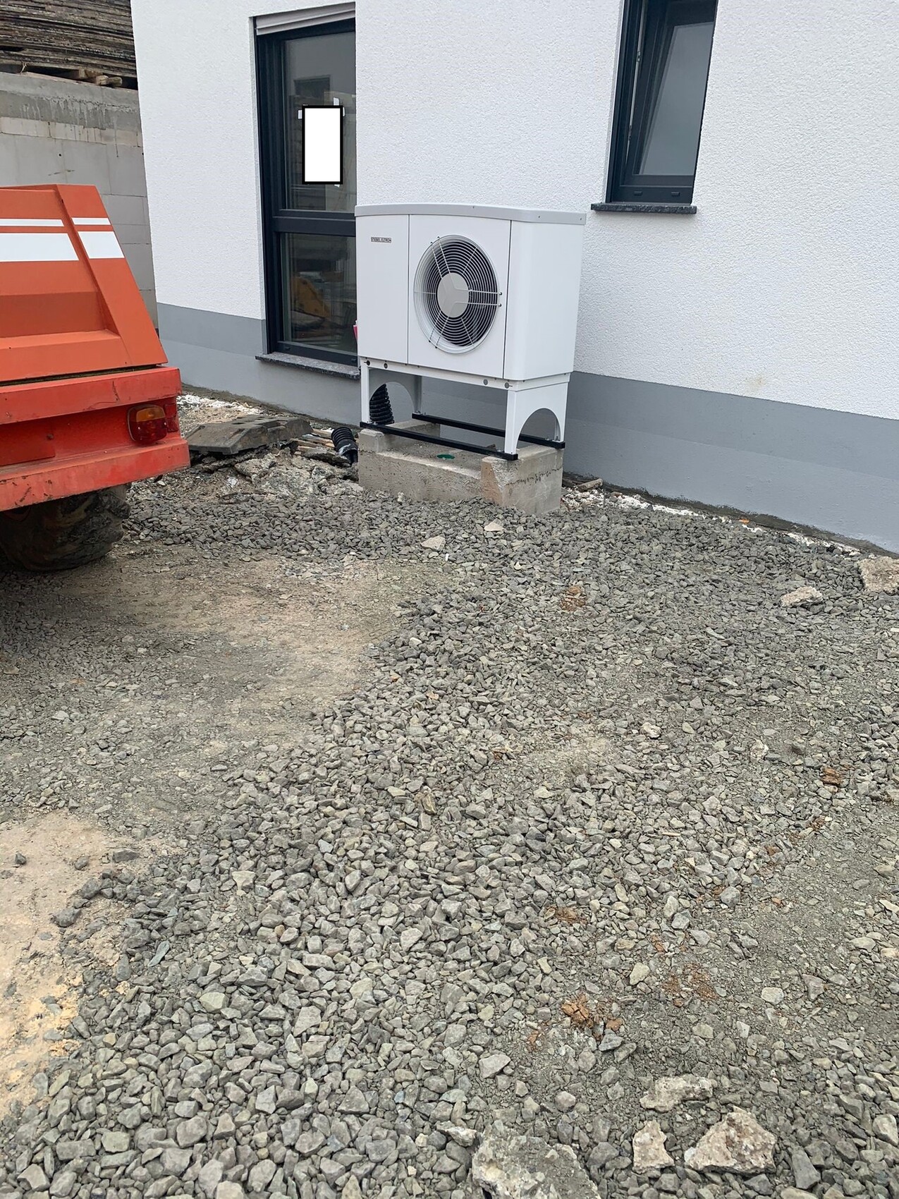 Outdoor area of a house with heat pump outdoor unit on a base in front of gravel ground