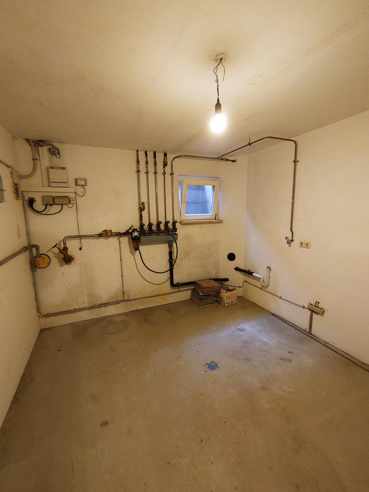 Basement room with heating and plumbing pipes, cables, light bulb, window, and floor drain.