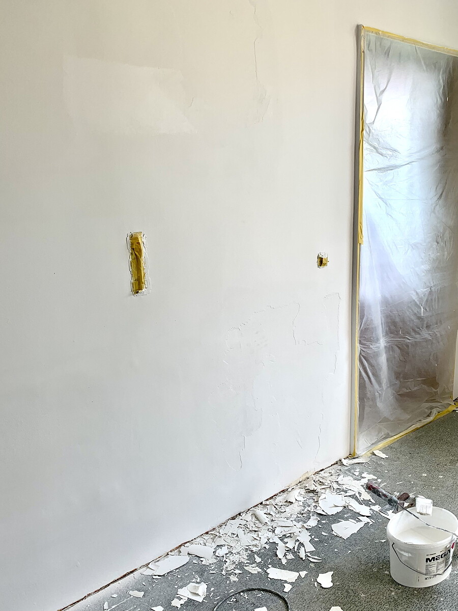 Renovation room: white wall with plaster cracks, door area behind plastic foil, floor with debris.