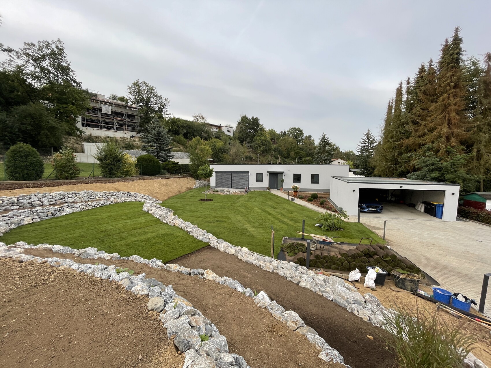 Modern housing complex with garage, green lawn, and stone terraces; construction work behind.