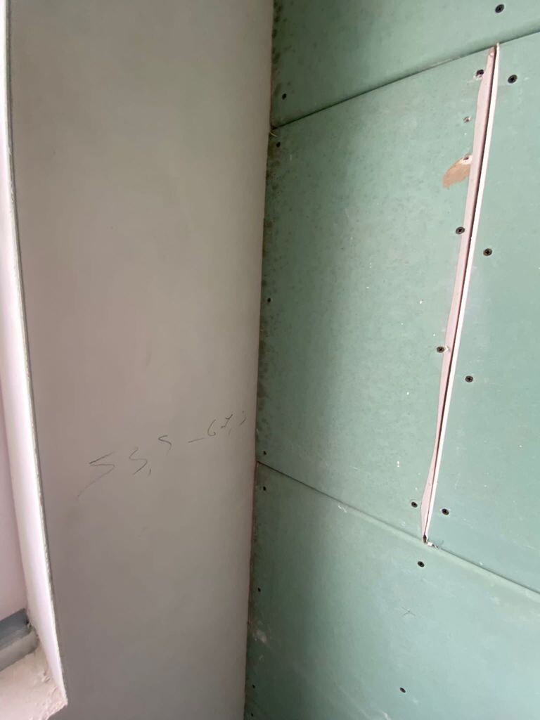 Interior: gray wall next to green drywall panels with screws and joint tape at the seam.