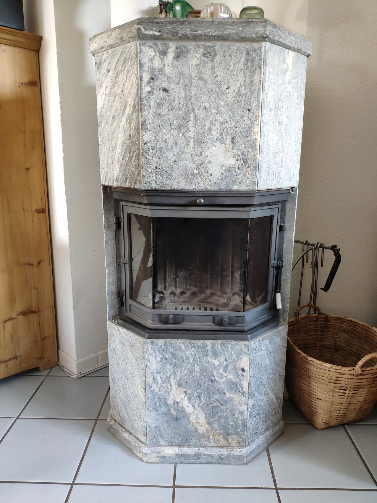 Freestanding wood-burning stove made of gray stone with glass front; next to it a wood basket.