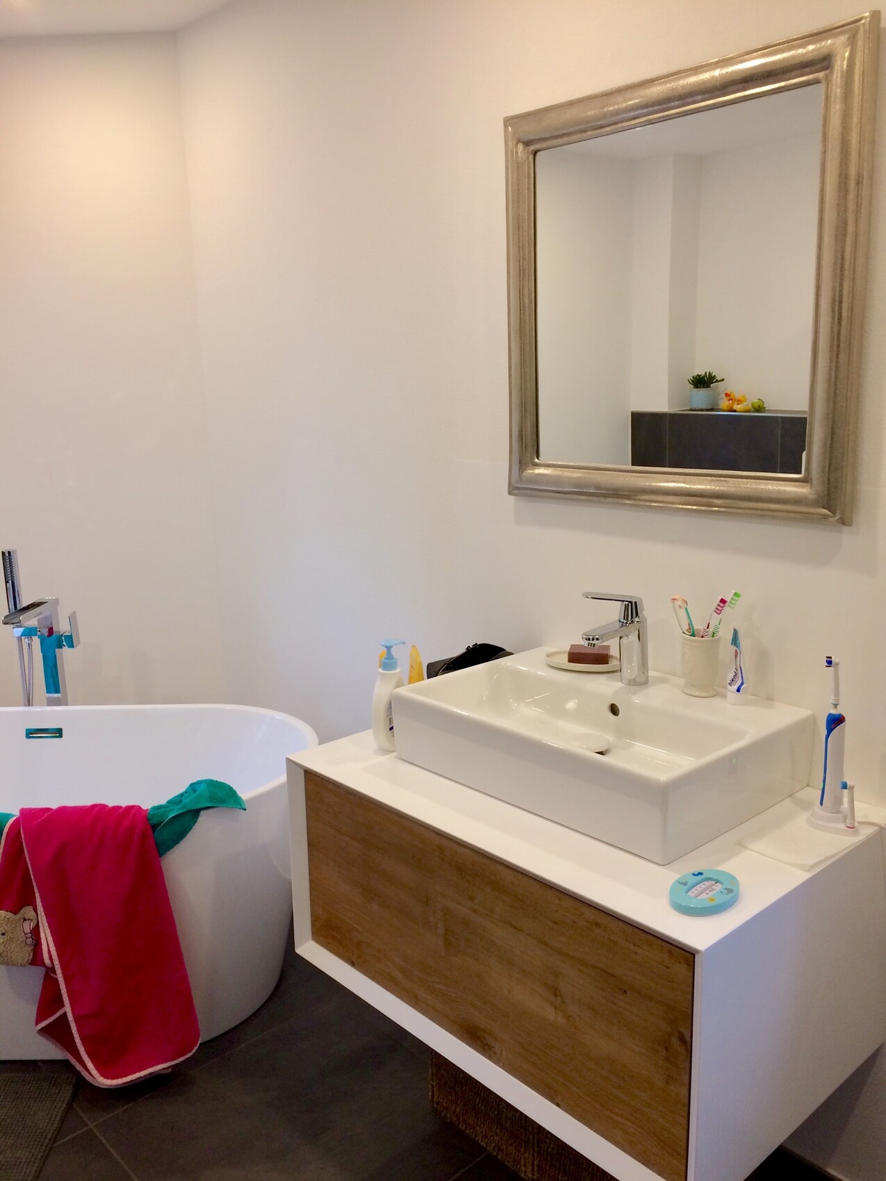 Modern bathroom: bathtub on the left, sink with wooden base, mirror frame, colorful towels.