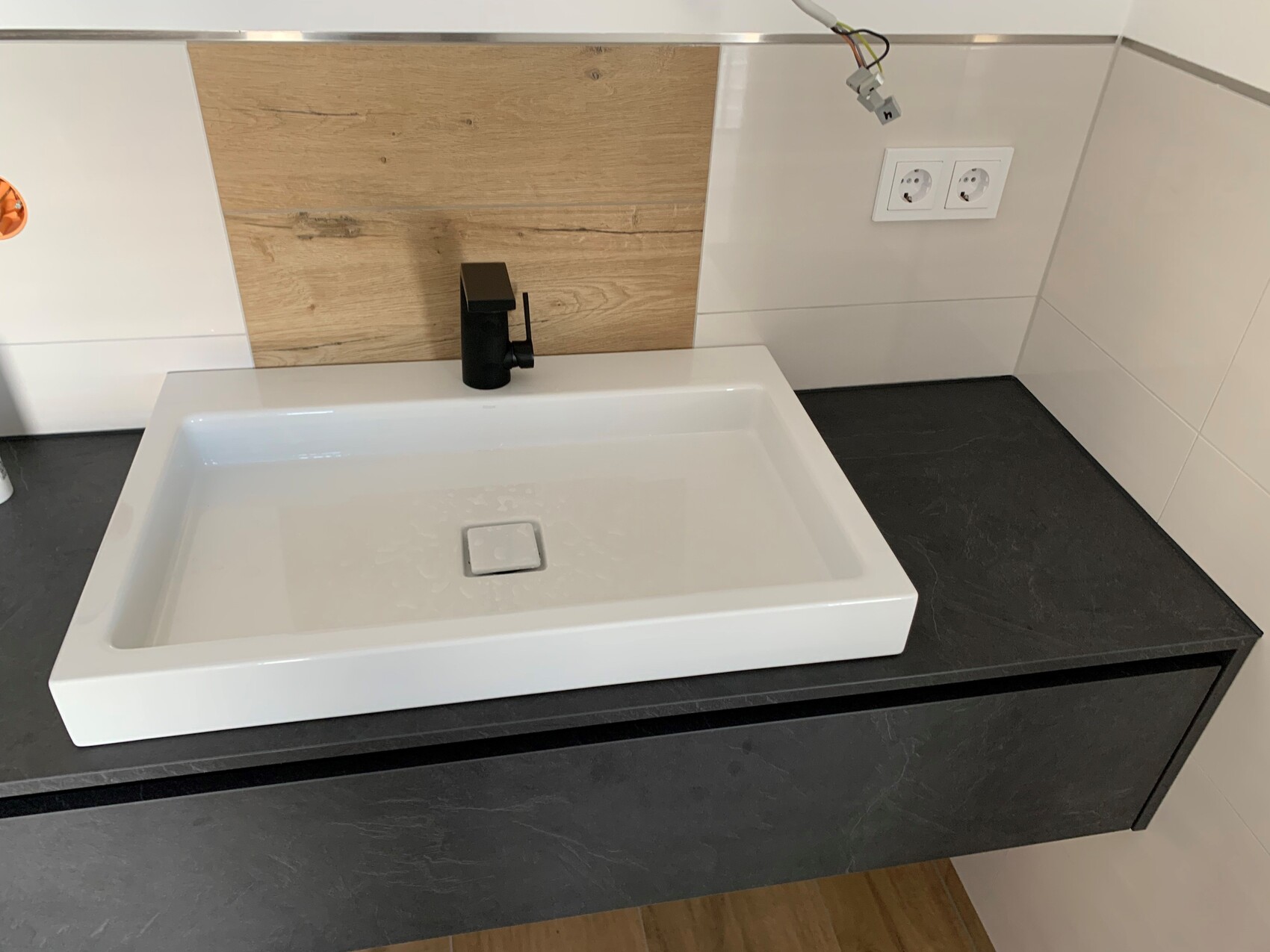 Rectangular white washbasin on dark countertop with black faucet, wooden backsplash.