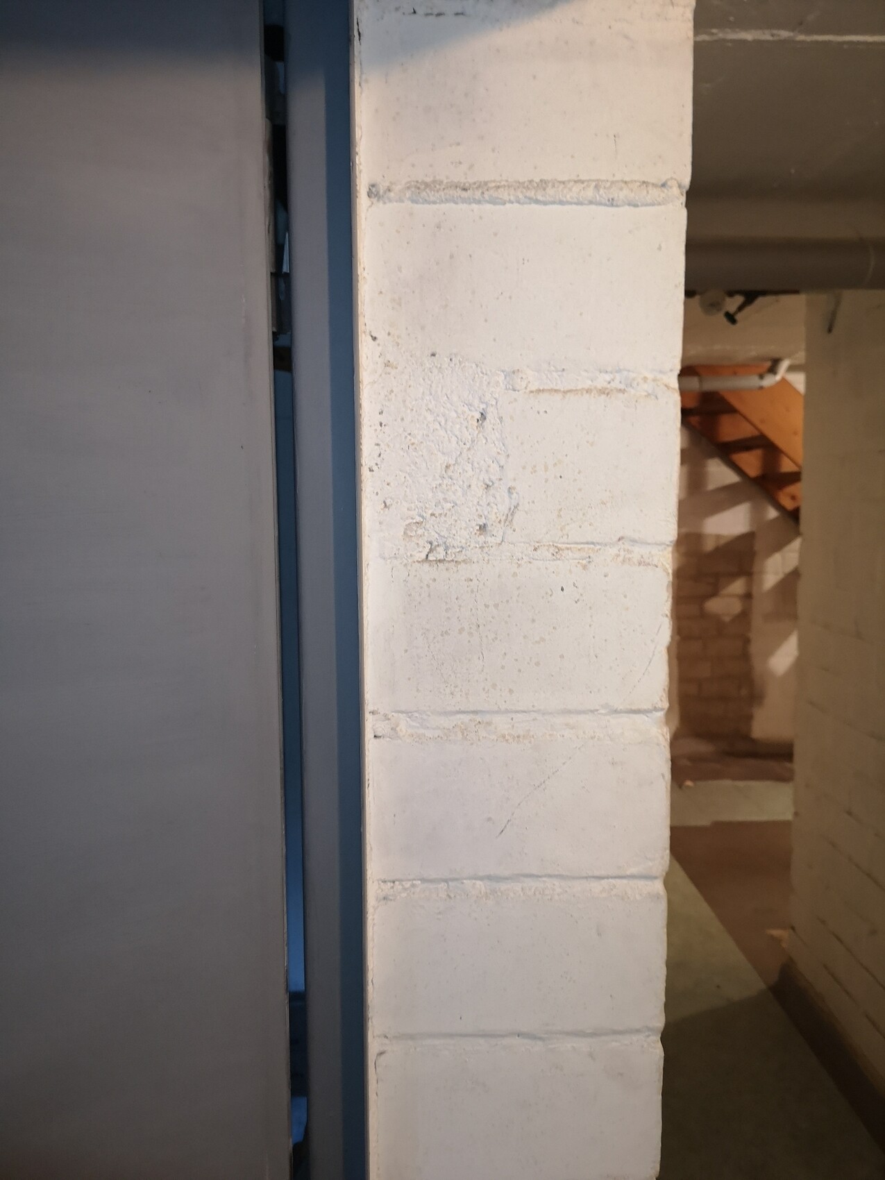 Mittig eine weiße Ziegelwand, links Metalltür, rechts Kellergang mit Holztreppe im Hintergrund.