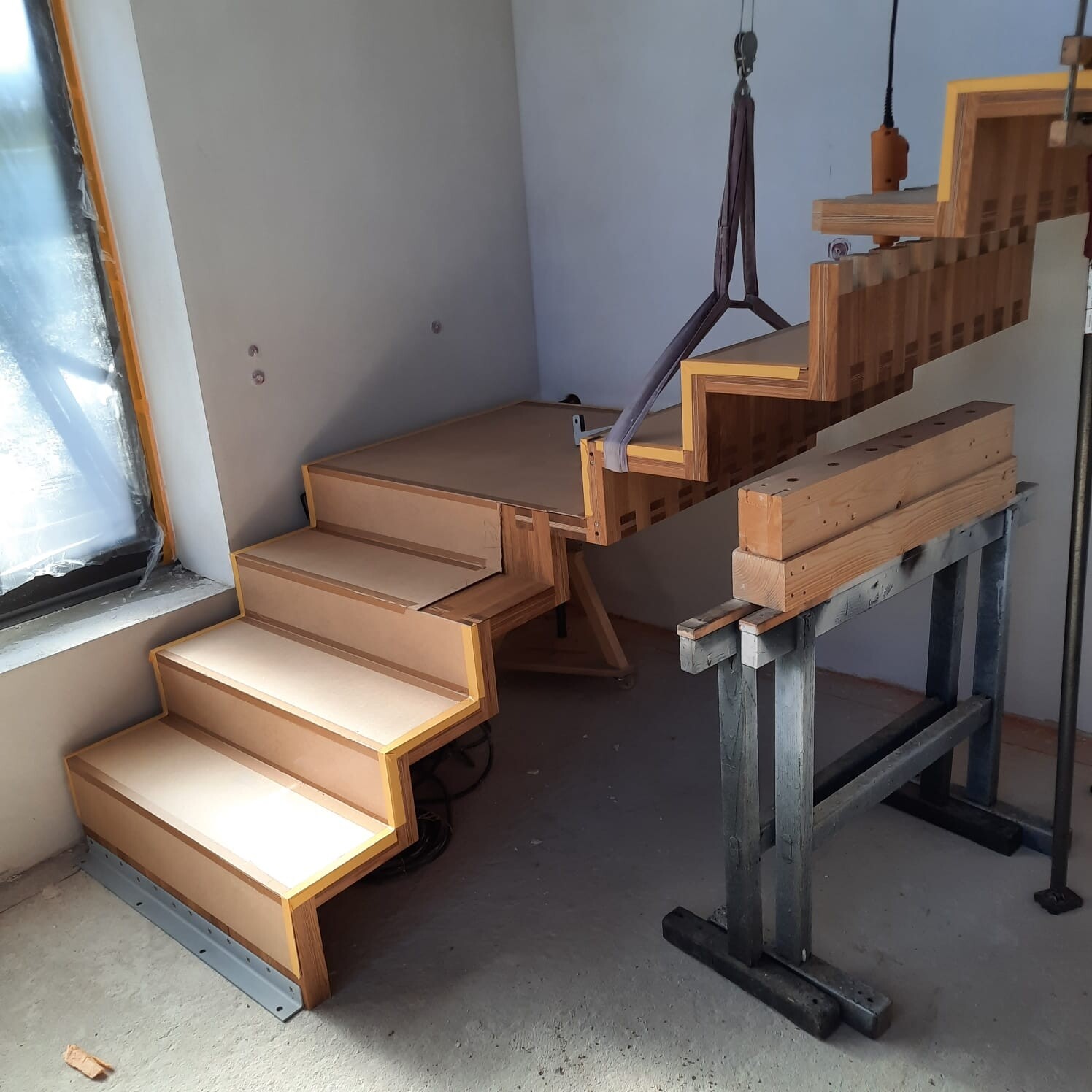 Partially assembled wooden staircase in workshop, crane strap hanging over the steps