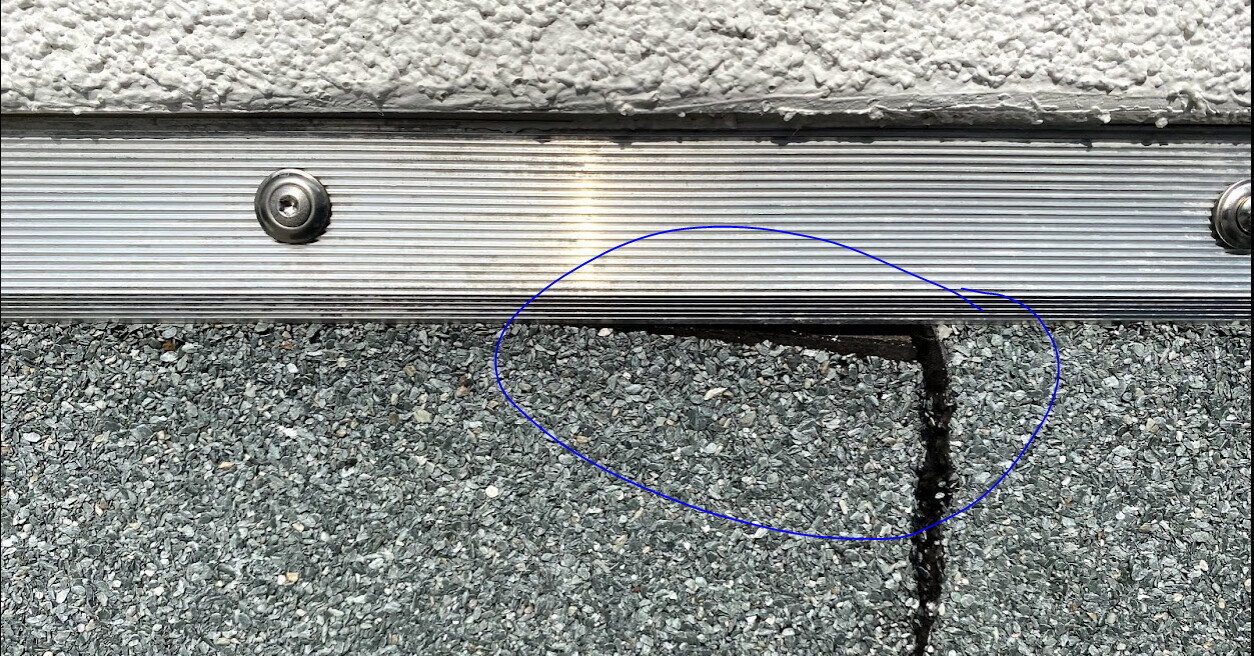 Metal threshold with grooves over coarse asphalt; rectangular gap at the lower edge.